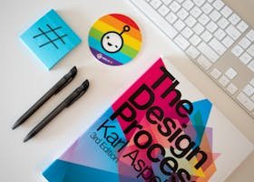 Flat lay of a creative desk setup featuring a design book, pens, and rainbow-themed sticker.